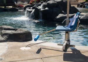 Chair lift for disabled person to enter resort swimming pool on hot sunny day with waterfall in the background